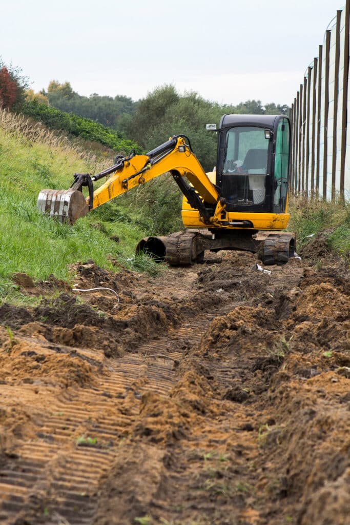 Une pelleteuse jaune est garée sur un terrain accidenté à Mont-de-Marsan, dans les Landes, à côté d'une pente herbeuse et d'une haute clôture, son bras étendu au-dessus d'un chemin de terre et de boue contenant des débris de construction provenant de récents travaux de nivellement.