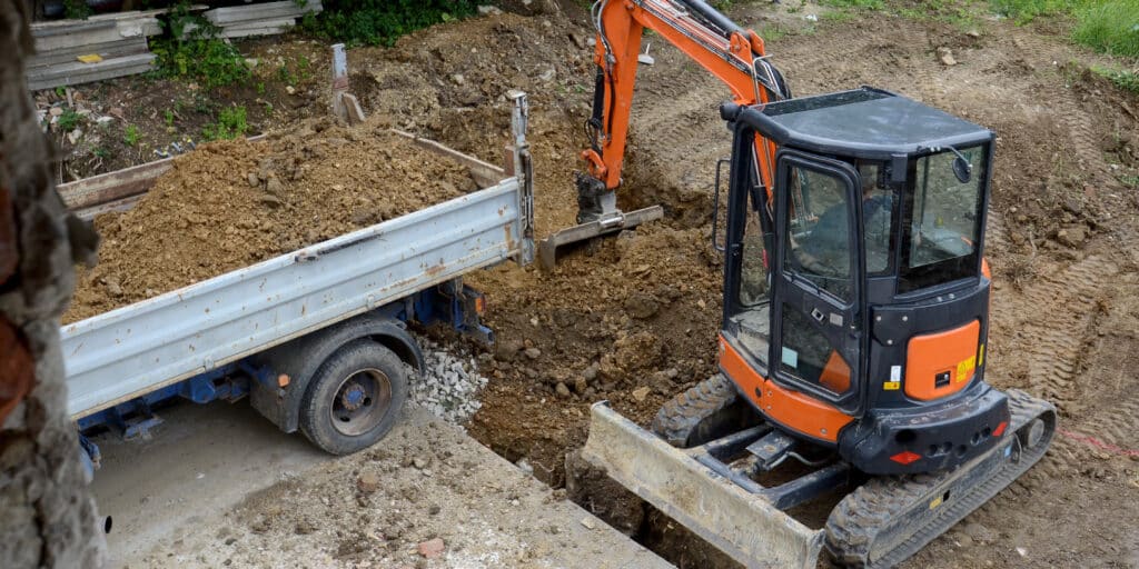 Une petite pelleteuse orange charge de la terre dans le plateau d'un camion-benne blanc sur un chantier de Mont-de-Marsan, où le terrassement et la terre nivelée se mélangent à des débris épars.
