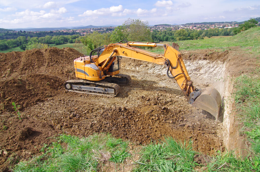 Une pelleteuse jaune effectue un terrassement, creusant un grand trou pour une fosse septique dans un champ de terre près de Mont-de-Marsan, avec de l'herbe verte et des arbres en arrière-plan sous un ciel partiellement nuageux.