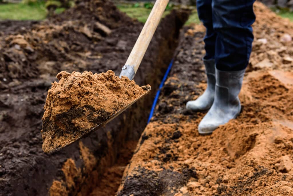 Une personne portant des bottes en caoutchouc se tient à côté d'une tranchée dans les Landes, tenant une pelle remplie de terre. L'image se concentre sur la pelle et la terre, mettant en évidence un travail typique de terrassement ou d'assainissement en cours.